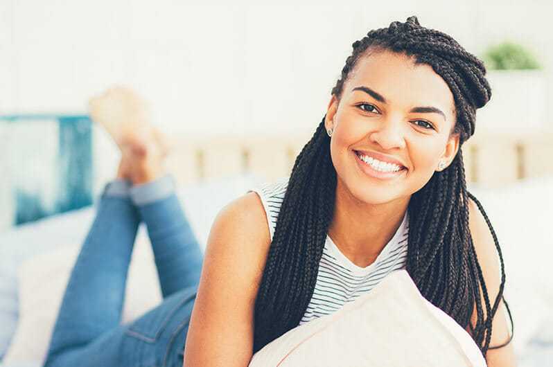 Smiling woman with braided hair, lying on a bed holding a pillow in a bright room.