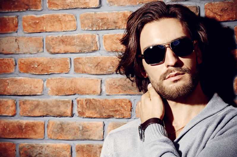 Man in sunglasses with beard poses against a brick wall.