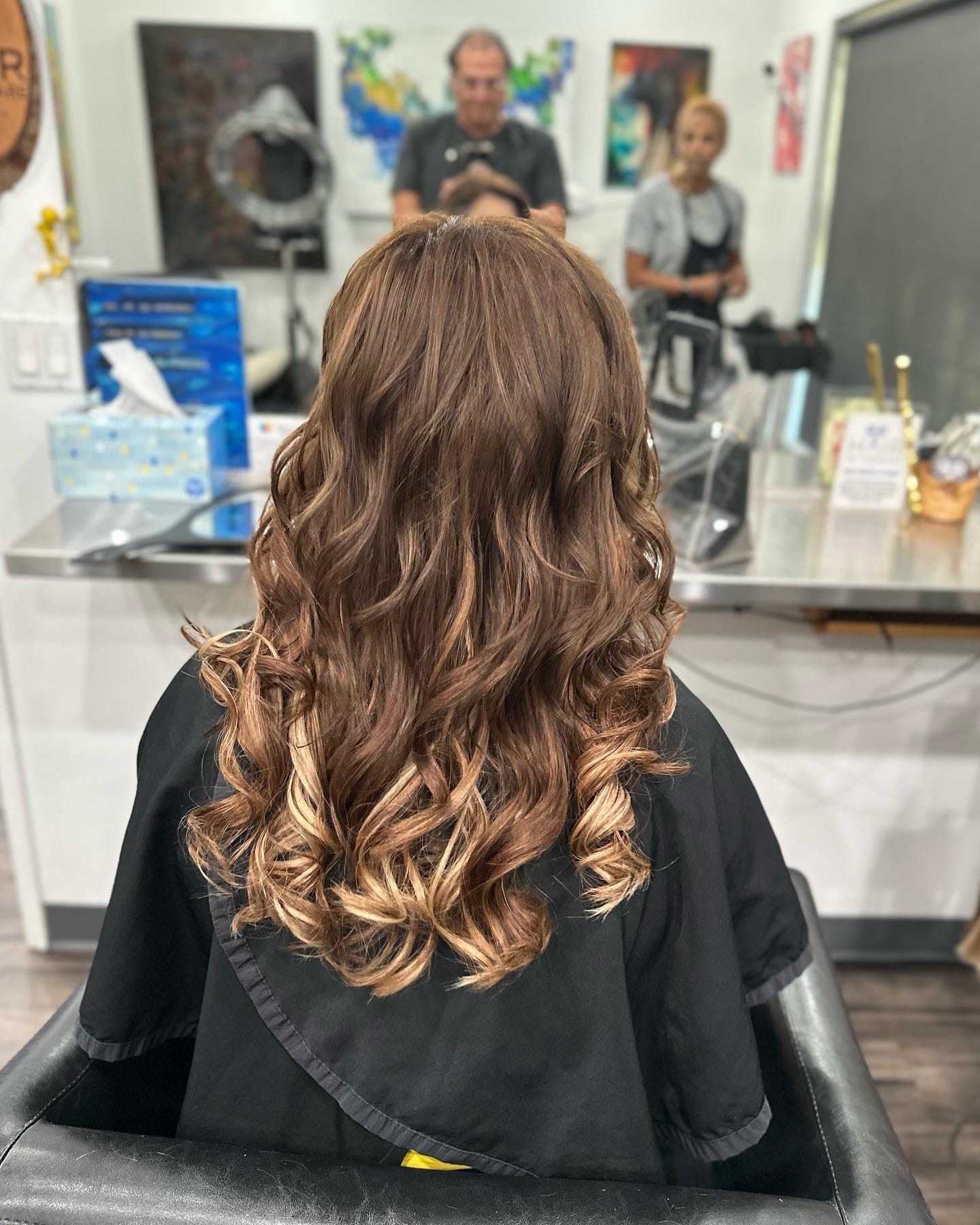 Woman with curled hair sitting in a salon chair, mirrors and stylists in the background.