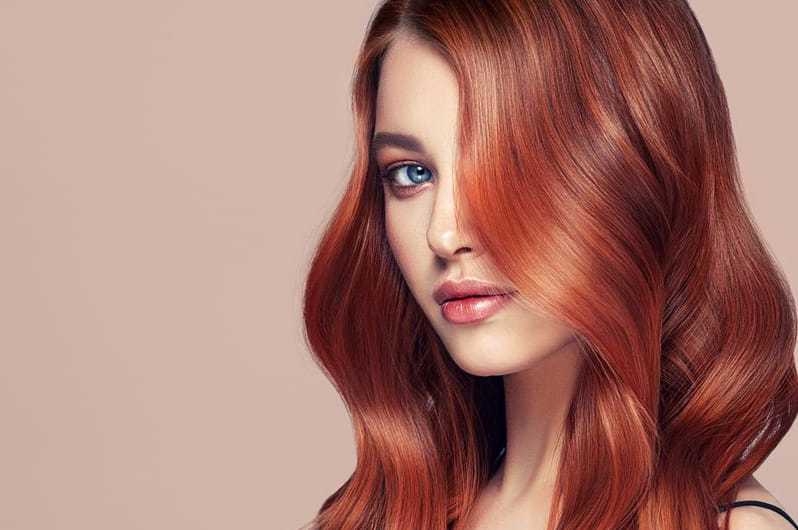 Young woman with long, wavy red hair covering one eye, against a beige background.
