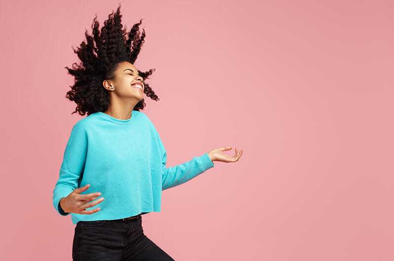 Woman in turquoise sweater joyfully jumping with hair flying against a pink background.